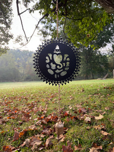 Decorative wind chime with Ganesha design hanging in a park with grass and trees.